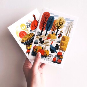 K&ouml;nnte beinhalten: Zwei illustrierte Karten mit Thanksgiving-Motiven. Eine Karte zeigt eine gebratene Pute und einen Kuchen. Die andere Karte zeigt eine Herbstlandschaft mit K&uuml;rbissen, Truth&uuml;hnern, Heuballen und einer Vogelscheuche. Die Karten werden von einer Person gehalten.