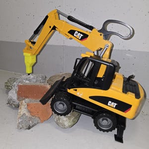 Pode incluir: Uma escavadora de brinquedo amarela com uma cabine preta e um martelo pneumático preto e amarelo. A escavadora está a quebrar um monte de tijolos.