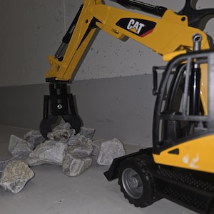 Op de afbeelding: Een gele speelgoedgraafmachine met een zwarte grijper, met het label "CAT", pakt grijze stenen op.