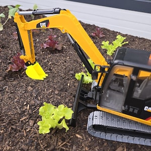 Könnte beinhalten: Ein gelber und schwarzer Spielzeugbagger mit dem CAT-Logo auf Arm und Kabine. Der Bagger befindet sich in einem Gartenbeet mit grünen und roten Salatpflanzen. Die Schaufel des Baggers ist gelb.