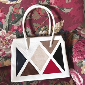 May include: A white handbag featuring a geometric pattern of triangles and a central diamond shape. The design incorporates panels in navy blue, brown, beige, black, and red. The bag has two white top handles and a structured rectangular silhouette.