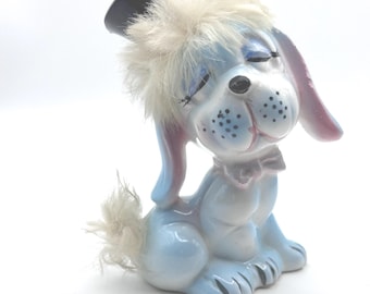 May include: A ceramic dog figurine with a glossy light blue finish, featuring closed eyes with painted eyelashes, a black nose, and a small bow at the neck. The dog wears a black top hat adorned with a tuft of white faux fur, matching its tail.