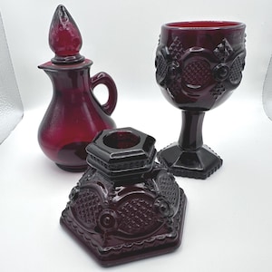 May include: A collection of ruby red glass items, including a decanter with a stopper, a goblet, and a decorative hexagonal piece. The pieces have embossed floral and geometric patterns, creating a vintage aesthetic.