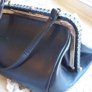 May include: A vintage black handbag with a structured design. The purse features a top handle, a metal clasp, and decorative trim along the frame. The bag appears to be made of leather or a similar material.