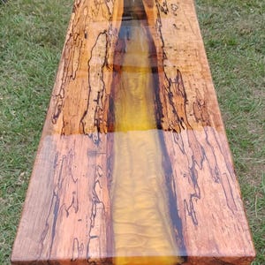 May include: A handcrafted wooden bench with a live edge design. The bench features a river of golden epoxy resin running through the center, set on black metal legs. The wood has a natural, warm tone with dark grain patterns.