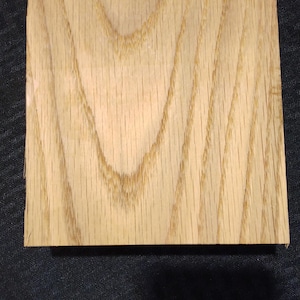 May include: A square, light-colored wooden block with visible grain patterns. The wood has a natural, warm tone, and the grain creates a textured appearance. The block is likely a sample or a small piece of lumber.