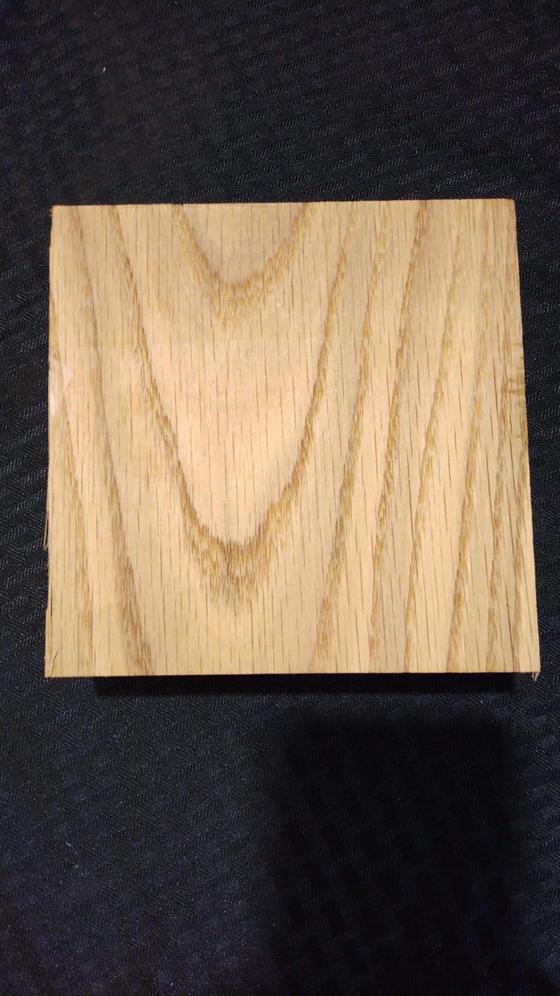 May include: A square, light-colored wooden block with visible grain patterns. The wood has a natural, warm tone, and the grain creates a textured appearance. The block is likely a sample or a small piece of lumber.