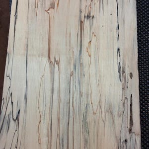 May include: A close-up of a piece of wood with a light beige base color and dark brown and black streaks. The wood grain pattern is visible, with a mix of straight and wavy lines. The wood appears to be a hardwood.