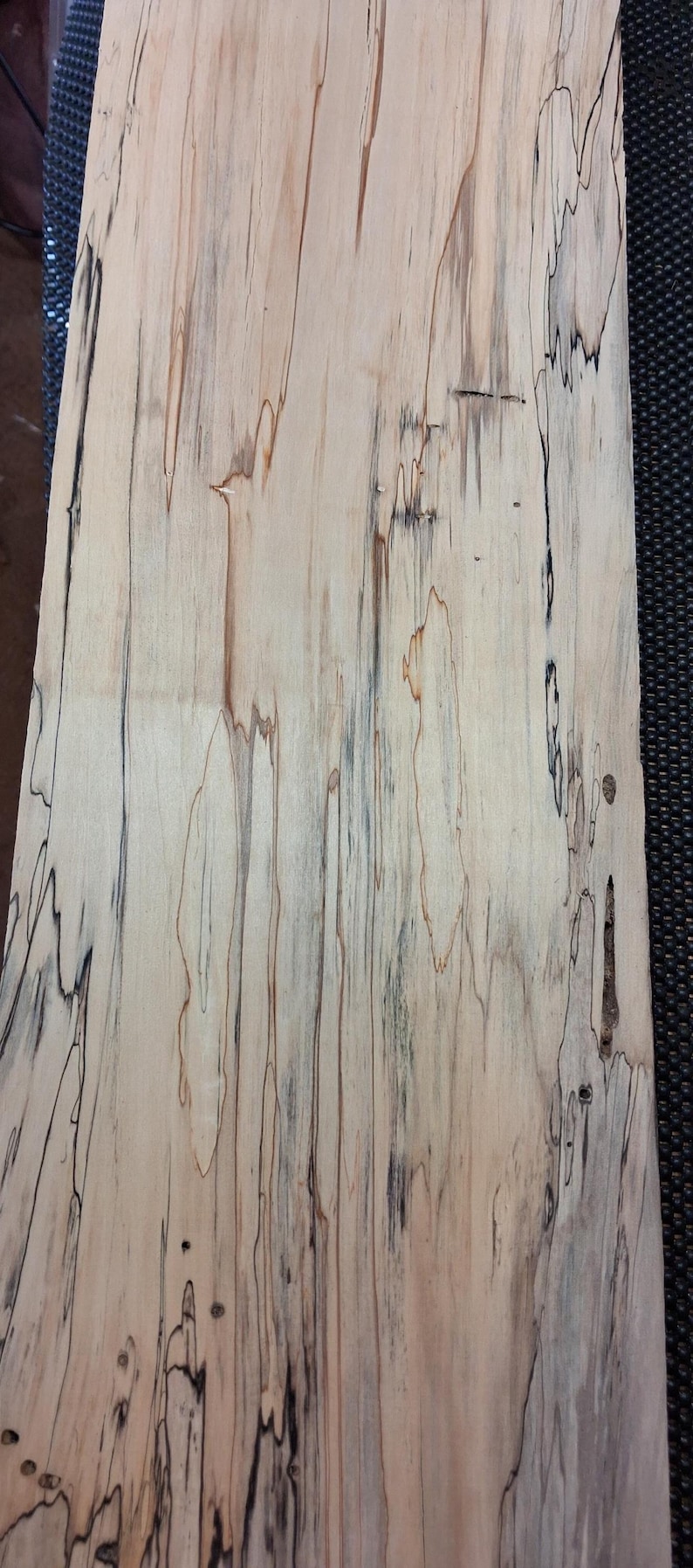 May include: A close-up of a piece of wood with a light beige base color and dark brown and black streaks. The wood grain pattern is visible, with a mix of straight and wavy lines. The wood appears to be a hardwood.
