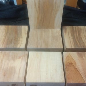 May include: A wooden chair in the process of being built, with a seat and backrest, surrounded by six square wooden blocks. The wood has a light brown color with visible grain patterns, suggesting a natural finish. The chair is unfinished.