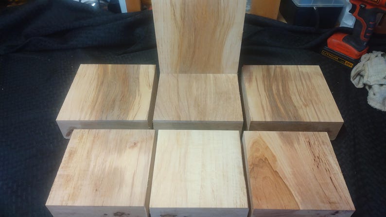 May include: A wooden chair in the process of being built, with a seat and backrest, surrounded by six square wooden blocks. The wood has a light brown color with visible grain patterns, suggesting a natural finish. The chair is unfinished.
