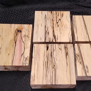 May include: A collection of six unfinished wood blocks. The blocks are rectangular, with a light tan color and striking black and reddish-brown grain patterns. The wood appears to be freshly cut and ready for crafting.