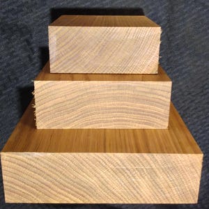 May include: Three stacked, rectangular wooden blocks with visible grain patterns. The blocks are a warm, light brown color, with the largest block at the base and the smallest on top. The wood appears to be unfinished.
