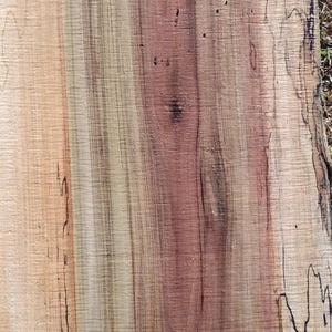 May include: Close-up of a wood slab with a natural grain pattern. The wood features a mix of light tan, reddish-brown, and dark brown hues, with dark streaks and knots. The texture is rough and organic.