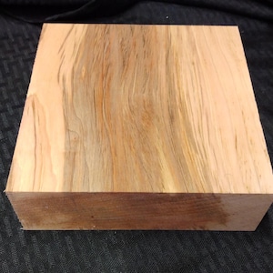May include: A square wooden block with a natural wood grain pattern. The wood features shades of light brown, tan, and darker brown, creating a visually appealing texture. The block is likely for woodworking or crafting projects.