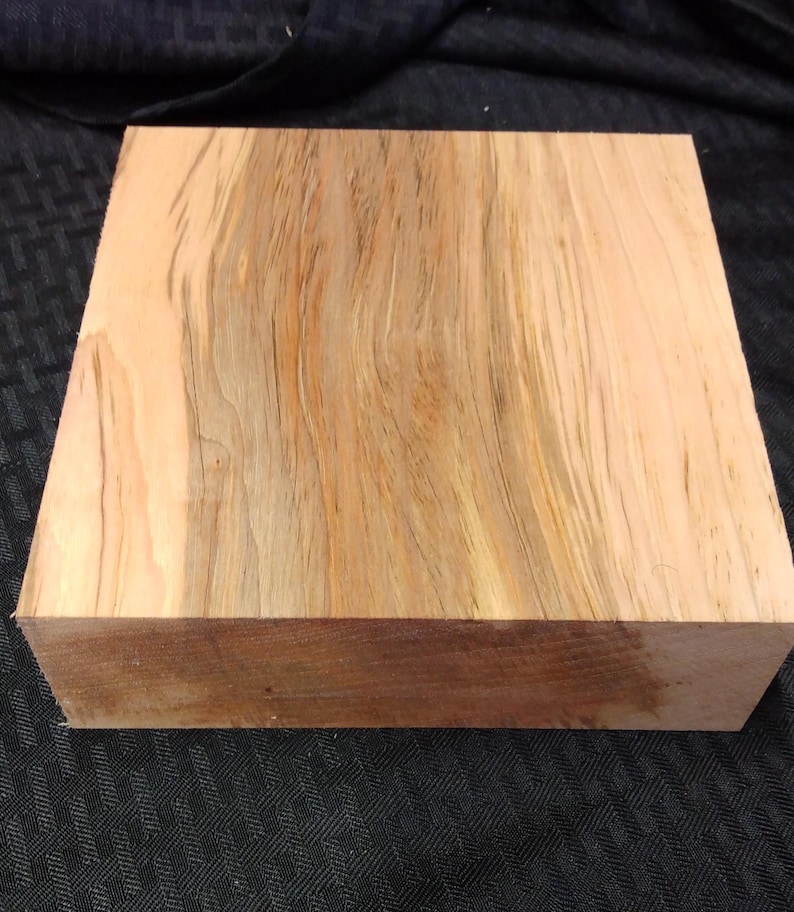 May include: A square wooden block with a natural wood grain pattern. The wood features shades of light brown, tan, and darker brown, creating a visually appealing texture. The block is likely for woodworking or crafting projects.