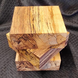 May include: Three stacked, unfinished wood blocks with a natural, light brown and tan color. The wood grain features dark brown and black streaks, creating a unique pattern. The blocks are cut into a rectangular shape, showcasing the wood's natural beauty.