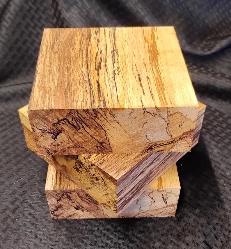 May include: Three stacked, unfinished wood blocks with a natural, light brown and tan color. The wood grain features dark brown and black streaks, creating a unique pattern. The blocks are cut into a rectangular shape, showcasing the wood's natural beauty.