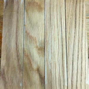 May include: Four unfinished wooden planks with a light brown color and visible wood grain. The planks are arranged side by side, showing the natural texture and color variations of the wood. The planks are likely for a woodworking project.