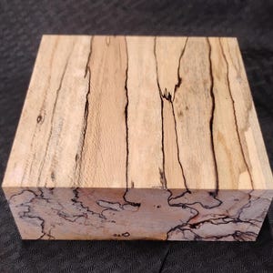 May include: A rectangular block of light-colored wood with dark, irregular grain patterns. The wood has a natural, unfinished appearance, showcasing the unique veining and color variations. The block is likely intended for woodworking or crafting projects.