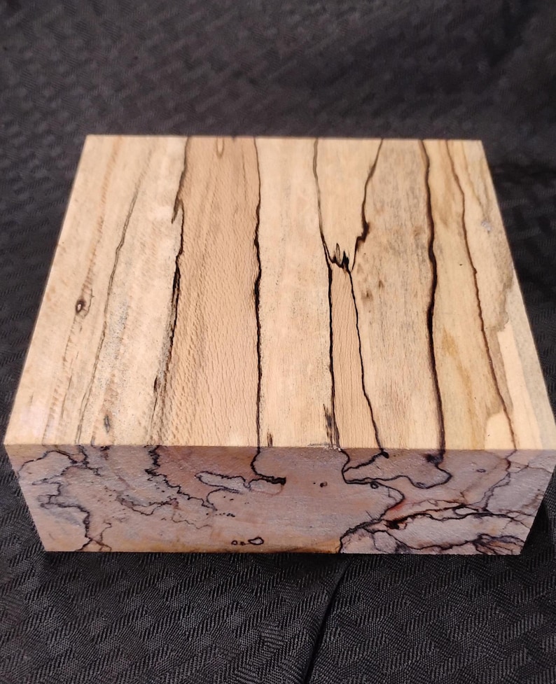 May include: A rectangular block of light-colored wood with dark, irregular grain patterns. The wood has a natural, unfinished appearance, showcasing the unique veining and color variations. The block is likely intended for woodworking or crafting projects.