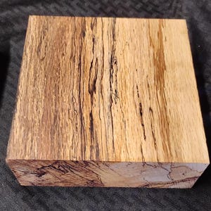May include: A rectangular block of wood with a natural, multi-toned grain pattern. The wood features shades of light brown and tan, with dark brown and black streaks running throughout. The block is likely for woodworking or crafting.
