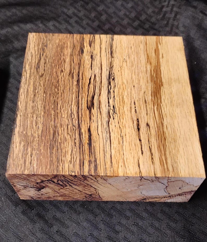 May include: A rectangular block of wood with a natural, multi-toned grain pattern. The wood features shades of light brown and tan, with dark brown and black streaks running throughout. The block is likely for woodworking or crafting.