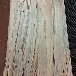 May include: A close-up of a piece of wood with a unique grain pattern. The wood has a light beige base color with dark brown and black streaks running vertically. There are also small, dark holes scattered across the surface.