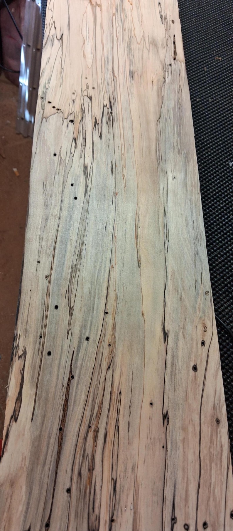 May include: A close-up of a piece of wood with a unique grain pattern. The wood has a light beige base color with dark brown and black streaks running vertically. There are also small, dark holes scattered across the surface.