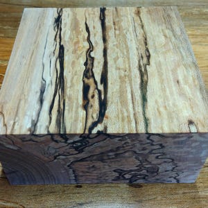 May include: A square wooden block with a unique grain pattern. The wood features a light tan base color with dark brown and black streaks running vertically and horizontally. The block's edges show a contrasting darker wood grain.