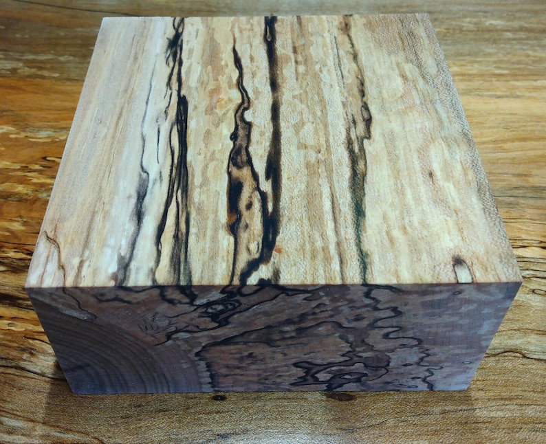 May include: A square wooden block with a unique grain pattern. The wood features a light tan base color with dark brown and black streaks running vertically and horizontally. The block's edges show a contrasting darker wood grain.