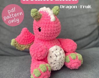 Puede incluir: Un juguete de dragón de crochet rosa con detalles verdes y un diseño de fruta de dragón de crochet blanco. El juguete está sentado en un sofá gris. El texto "Dragon - Fruit" y "pdf pattern only" es visible en la imagen.