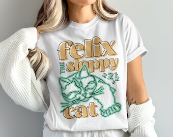 Peut inclure: T-shirt blanc avec un motif graphique d'un chat avec le texte "Felix the Sloppy Cat" en jaune et vert.