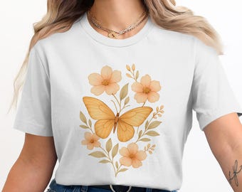 Peut inclure: T-shirt blanc avec un motif aquarelle d'un papillon et de fleurs aux tons orange et pêche. Le papillon est au centre, entouré de fleurs et de feuilles. Le motif est centré sur le t-shirt.