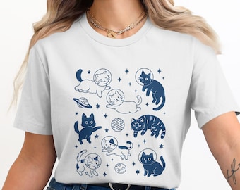 May include: White t-shirt featuring a navy blue graphic of cats in space helmets, planets, and stars. The design includes various cat poses and celestial elements, creating a whimsical, space-themed aesthetic.