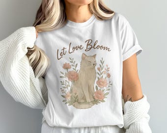 May include: White t-shirt featuring a watercolor illustration of a cat surrounded by pink flowers and green leaves. The text "Let Love Bloom" is written above the cat in a brown script font. The shirt is a casual, everyday wear item.