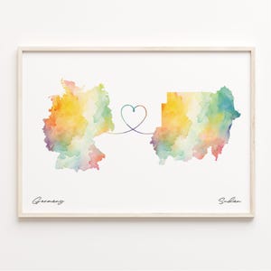May include: A framed watercolour print showing the maps of Germany and Sudan, linked by a heart-shaped line. The maps are filled with rainbow colours. The word "Germany" is written below the map of Germany, and "Sudan" is written below the map of Sudan.