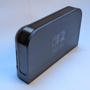 May include: A black Nintendo Switch dock with the Nintendo Switch logo and the number 2 printed on the front. The dock has rounded corners and a sleek, modern design.