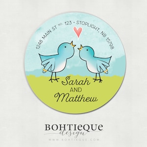 Op de afbeelding: Een ronde sticker met een groene achtergrond en twee blauwe vogels die elkaar aankijken. De vogels houden ringen vast. Boven de vogels bevindt zich een roze hart. De tekst "Sarah en Matthew" staat onder de vogels. De tekst "1248 Main St No. 123 Stoplight, NB 15798" staat rond de bovenkant van de sticker. De tekst "BOHTIEQUE design WWW BOHTIEQUE COM" staat onder de sticker.