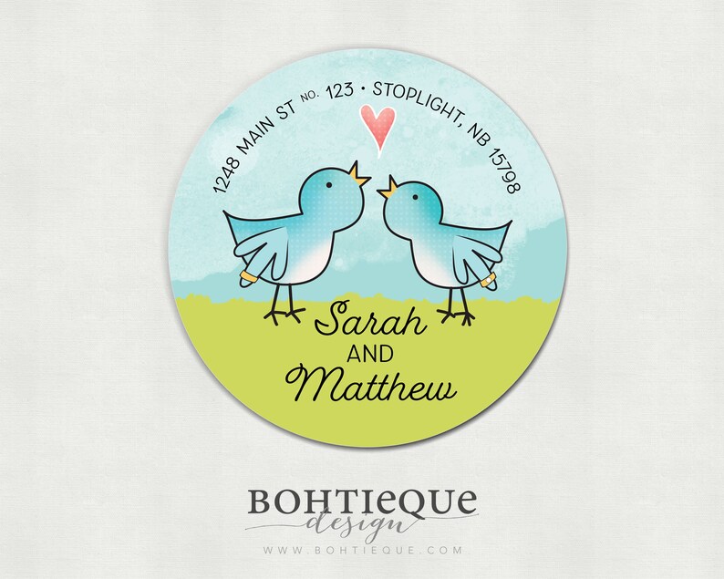 Op de afbeelding: Een ronde sticker met een groene achtergrond en twee blauwe vogels die elkaar aankijken. De vogels houden ringen vast. Boven de vogels bevindt zich een roze hart. De tekst "Sarah en Matthew" staat onder de vogels. De tekst "1248 Main St No. 123 Stoplight, NB 15798" staat rond de bovenkant van de sticker. De tekst "BOHTIEQUE design WWW BOHTIEQUE COM" staat onder de sticker.