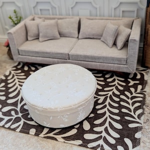 May include: Miniature living room set with a beige sofa, armchair, and a round ottoman on a brown and white patterned rug. A wooden cabinet with drawers is in the background. The scene is a dollhouse interior.