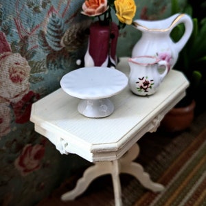 May include: A white porcelain cake stand with a scalloped edge sits on a white wooden table with a floral pattern. A white pitcher with gold trim and a smaller white pitcher with floral designs are also on the table. A vase with orange and yellow flowers is in the background.