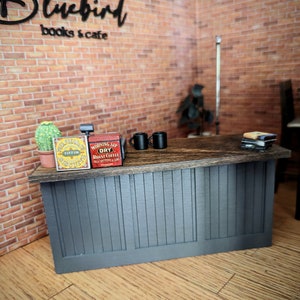 May include: Miniature cafe scene with a counter, featuring a sign that reads "Bluebird books & cafe." The counter has a wooden top, a gray front, and includes miniature items like coffee tins, mugs, a cactus, and books. The background is a brick wall.