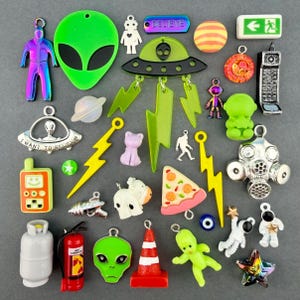 Op de afbeelding: Een verzameling diverse bedels, waaronder een groen alienhoofd, een UFO, een gasmasker, een brandblusser, een pizzapunt, een verkeerskegel, een neon astronaut, een gastank, een bliksemschicht, een robot en een bordje met een pijl en rennend figuur.
