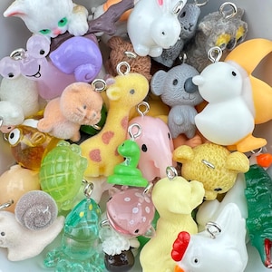 Puede incluir: Una colección de colgantes con forma de animales de colores, incluyendo una jirafa, un koala, un pato y un conejo. Los colgantes están hechos de varios materiales y tienen lazos plateados para sujetarlos a joyas u otras manualidades.