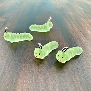 May include: Four translucent green caterpillar charms with silver-colored loops. Each caterpillar has a smiling face with black eyes and a red mouth. The charms are arranged on a dark wooden surface.
