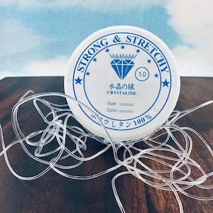 May include: A spool of clear, stretchy string with the text "STRONG & STRETCHY" in blue. The spool also has Japanese text and a diamond graphic. The string is 1.0 mm in diameter and is made of 100% polyurethane.