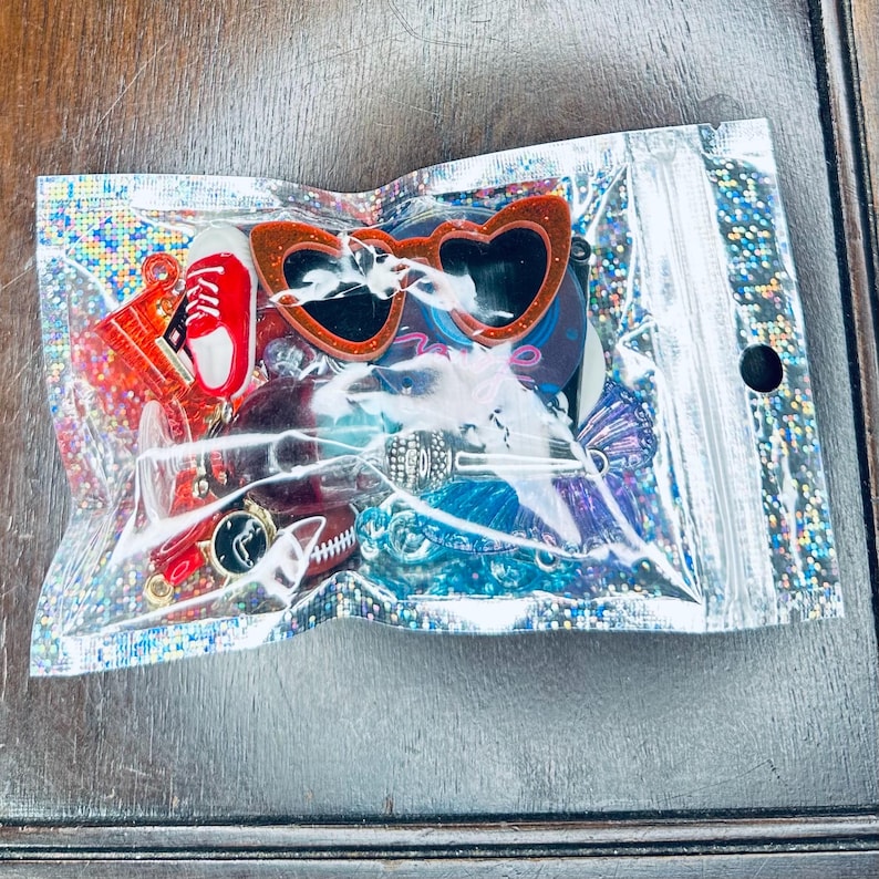 May include: A clear, iridescent bag filled with various charms and accessories. Notable items include red heart-shaped sunglasses, a red sneaker charm, and other colorful trinkets. The bag is sealed with a black dot.