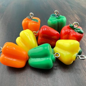 Puede incluir: Una colección de colgantes con forma de pimiento en colores naranja, verde, rojo y amarillo. Cada colgante tiene un lazo plateado para sujetarlo a joyas u otras manualidades. Los pimientos son brillantes y parecen estar hechos de resina.
