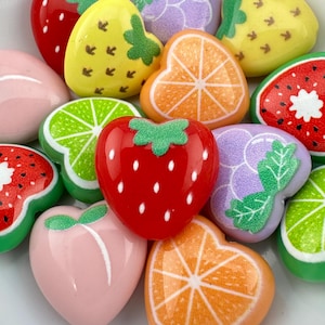 May include: A collection of colorful fruit-shaped embellishments, including strawberries, oranges, grapes, and watermelon. The embellishments are in various shades of red, yellow, green, and pink, with detailed designs and textures. These are ideal for crafting and DIY projects.